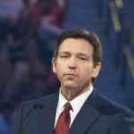 DeSantis Puts Trump Name on Airport Man in suit speaking at a public event.
