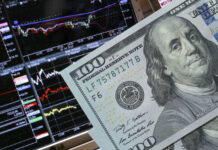 A close-up of a hundred-dollar bill in front of stock market graphs