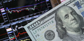 A close-up of a hundred-dollar bill in front of stock market graphs