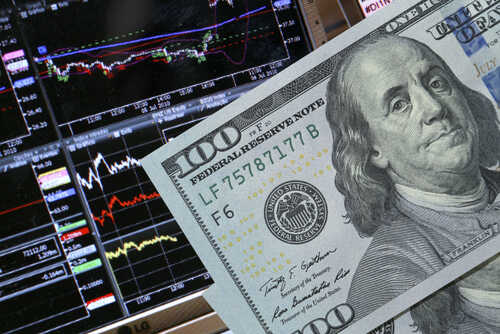 A close-up of a hundred-dollar bill in front of stock market graphs