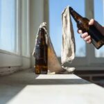 A hand holding a bottle next to another bottle covered with a cloth on a windowsill