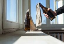 A hand holding a bottle next to another bottle covered with a cloth on a windowsill