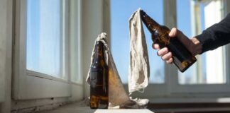 A hand holding a bottle next to another bottle covered with a cloth on a windowsill