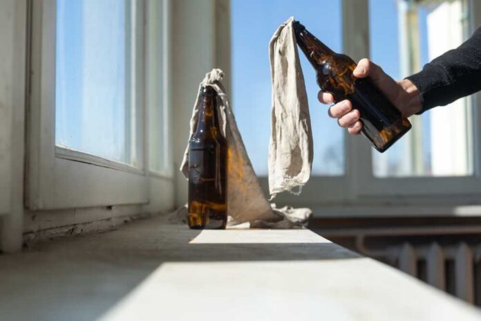 A hand holding a bottle next to another bottle covered with a cloth on a windowsill