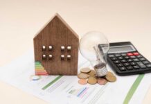 A wooden house model, light bulb, coins, and a calculator on financial documents