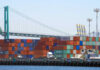 A view of stacked shipping containers at a port with a bridge in the background