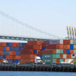 Food Backlog Explodes Amid Iran War A view of stacked shipping containers at a port with a bridge in the background