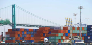 A view of stacked shipping containers at a port with a bridge in the background
