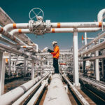 Deadly Blast Rocks Gas Facility Worker in orange jacket inspecting industrial pipes.
